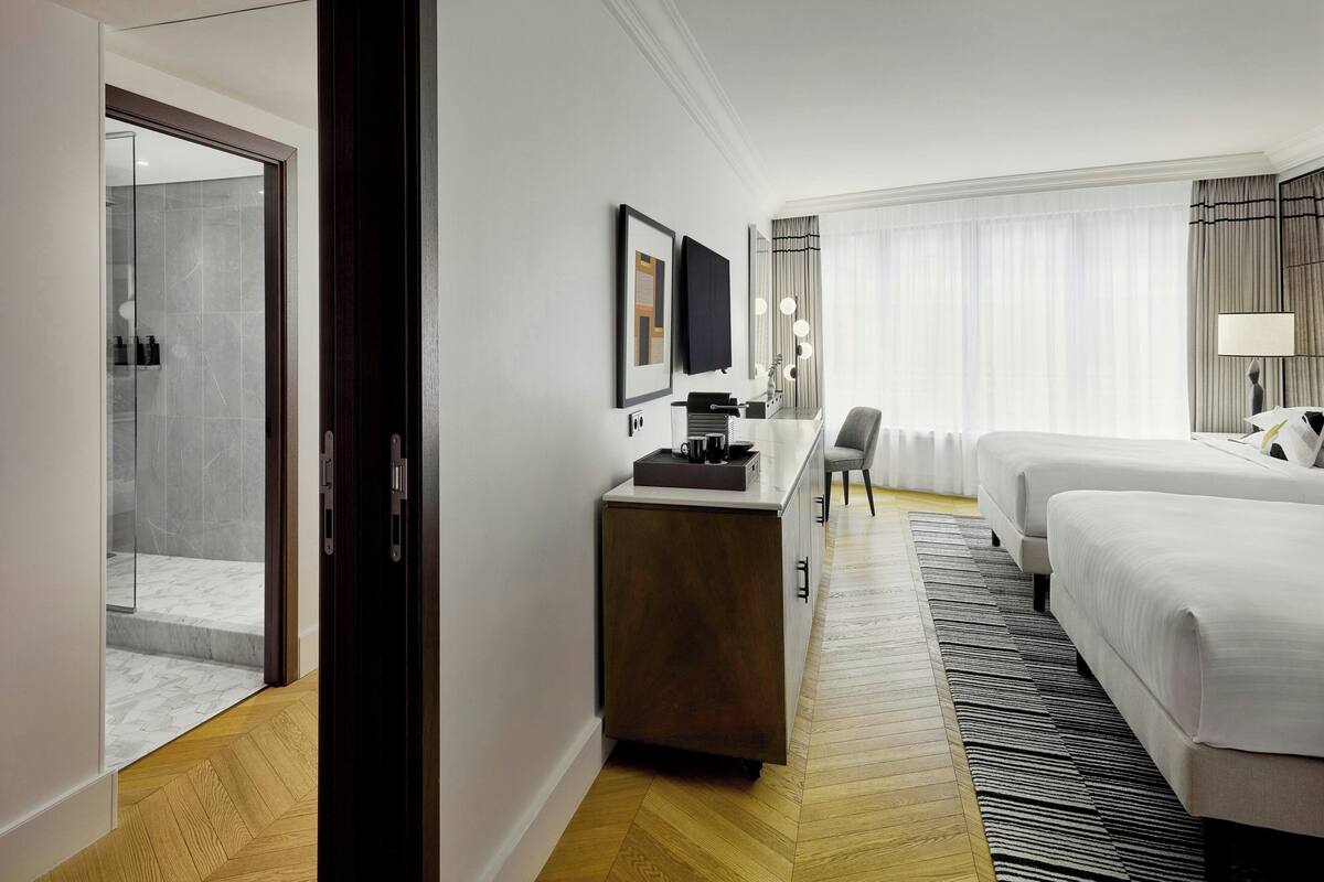 A hotel room with a bed, a desk with a computer, a chair, and a bathroom with a glass shower.