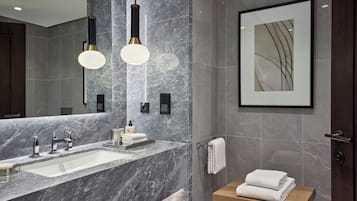 Un baño moderno con lavamanos de mármol, dos lámparas colgantes, una obra de arte enmarcada y un estante de madera con toallas.