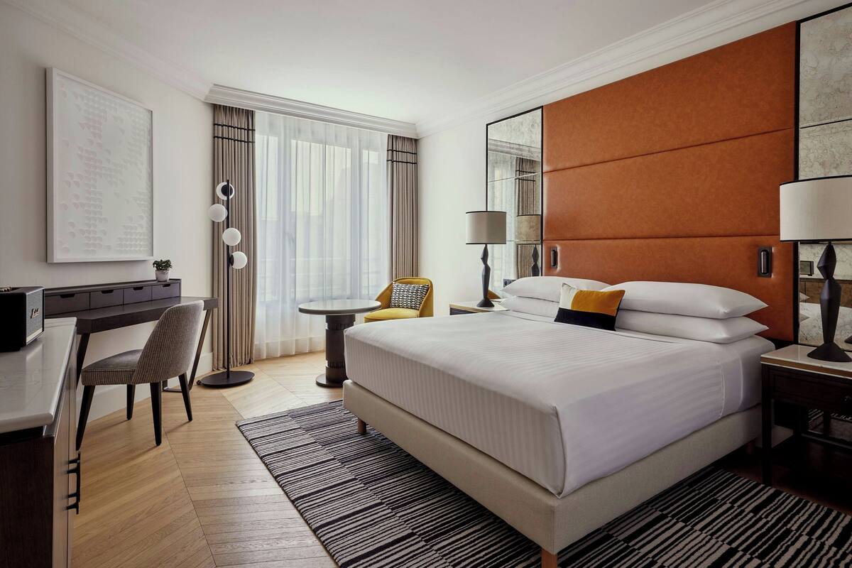 A hotel room with a large bed, a desk, a chair, a small table, and a lamp.
