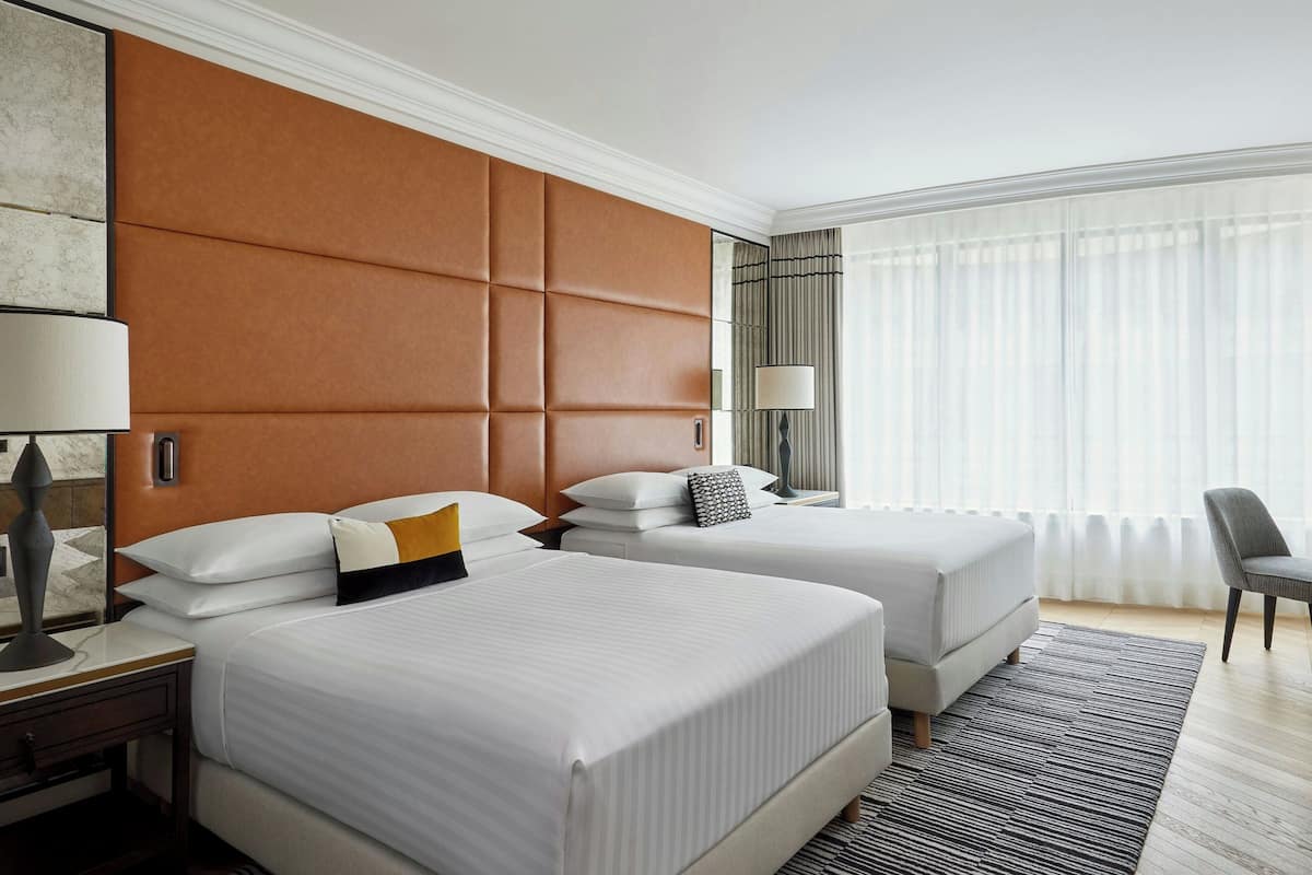 A hotel room with two beds, a large padded headboard, a desk with a lamp, and a chair.