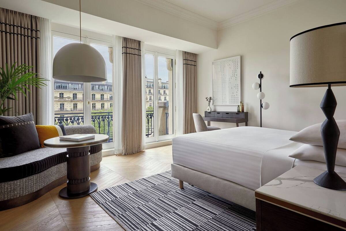 A hotel room with a large bed, a sofa, a dining table, and a balcony with a view of buildings.