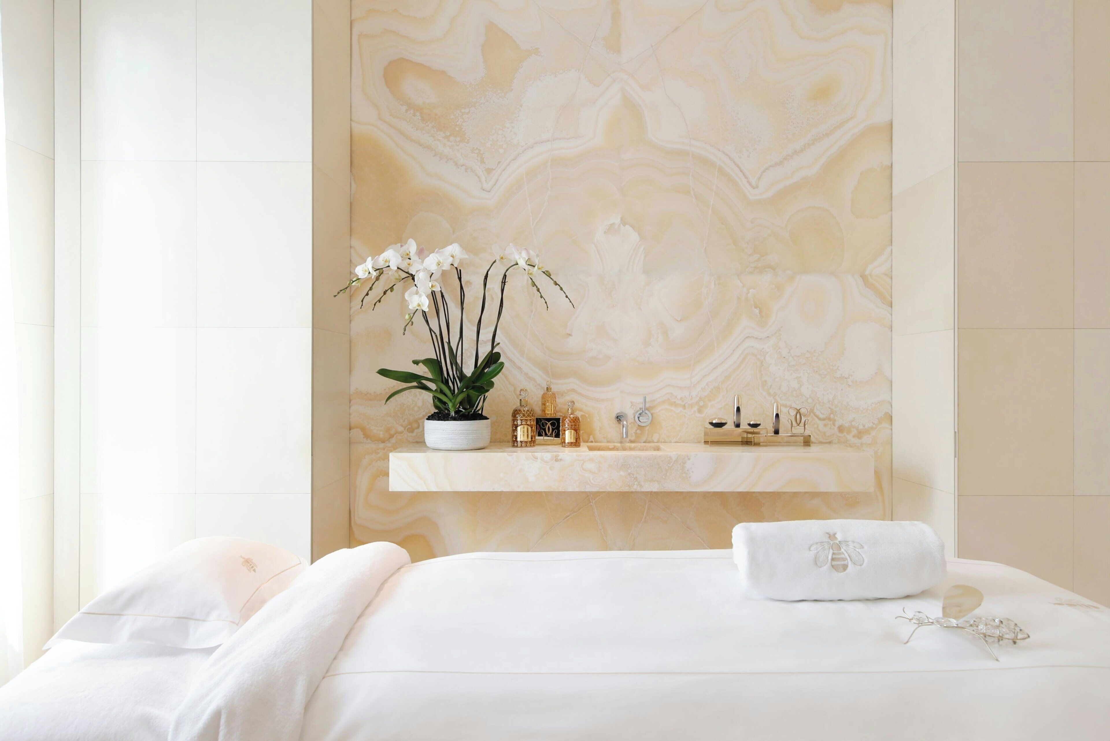 A hotel room with a marble wall, a white bed, a white bathrobe, a white towel, and a shelf with toiletries.