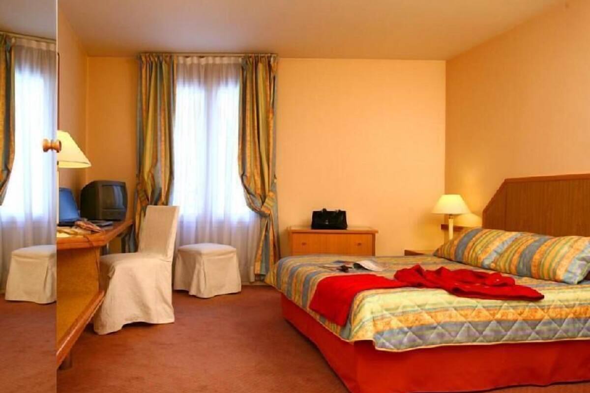 A hotel room with a bed, a desk with a computer, a chair, a television, and a nightstand with a lamp.