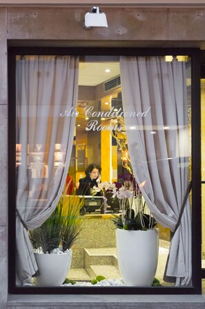 A hotel entrance with air-conditioned rooms, a woman in a black coat, and potted plants. - Hotel Fertel Etoile (Paris)