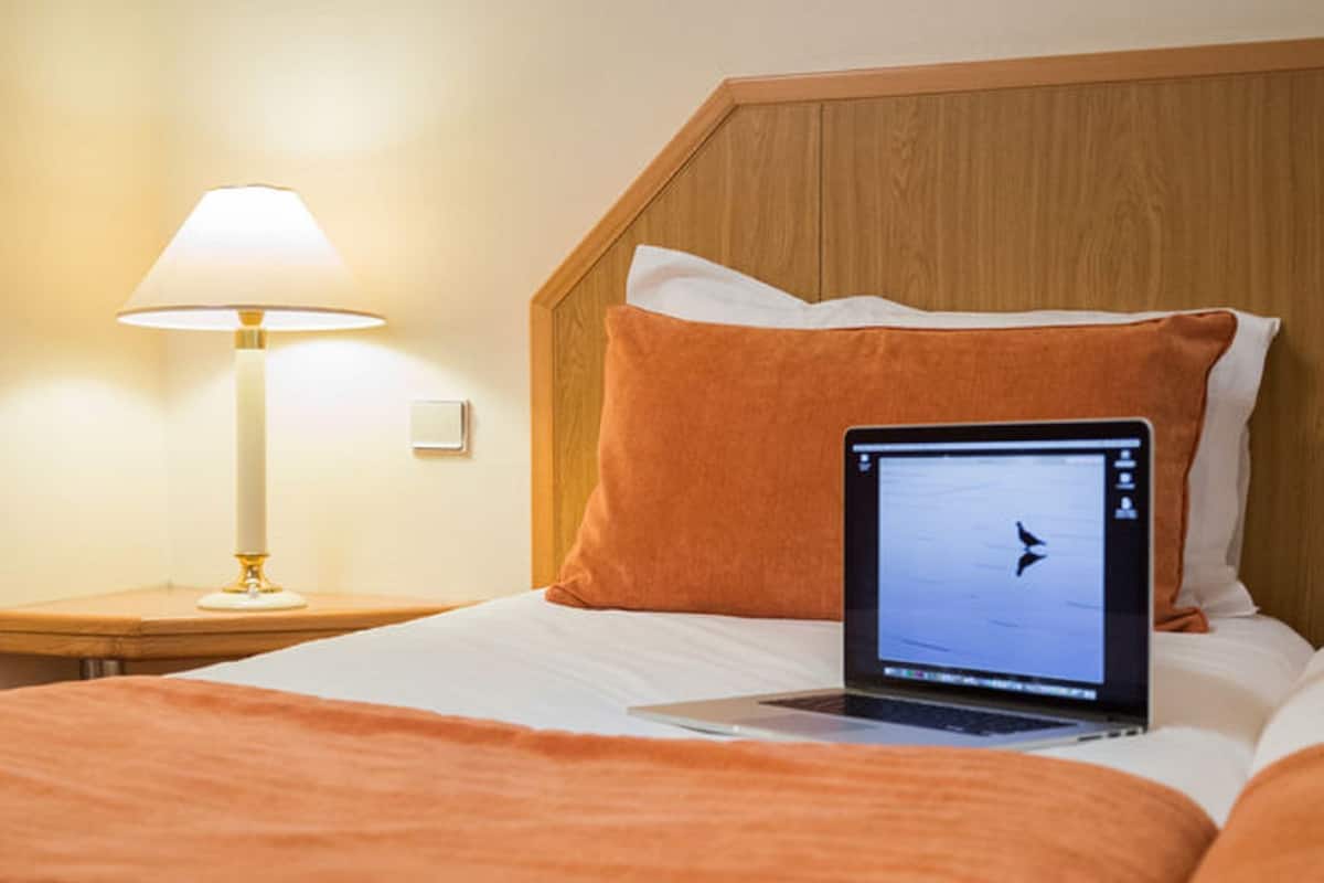 A neatly made bed with an orange blanket, a laptop on the bed, and a lamp on a bedside table.