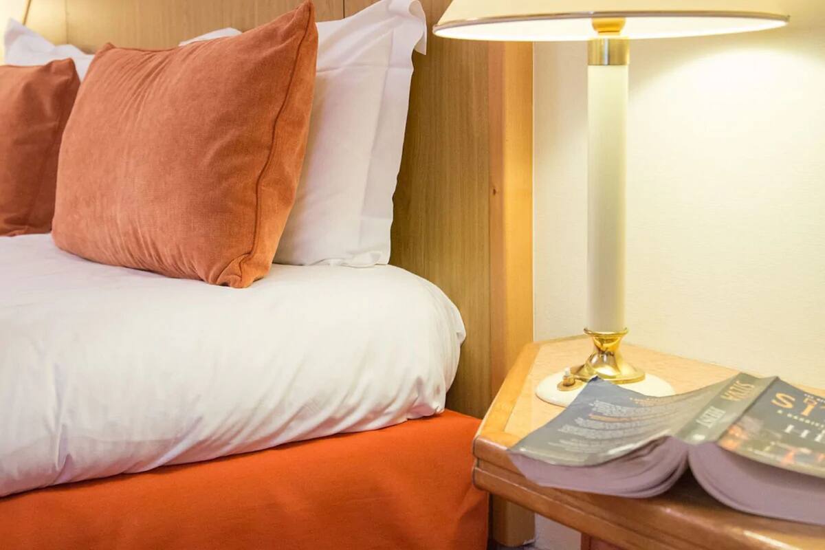 A neatly made bed with white and orange bedding, a wooden headboard, and a bedside table with a lamp and a book.
