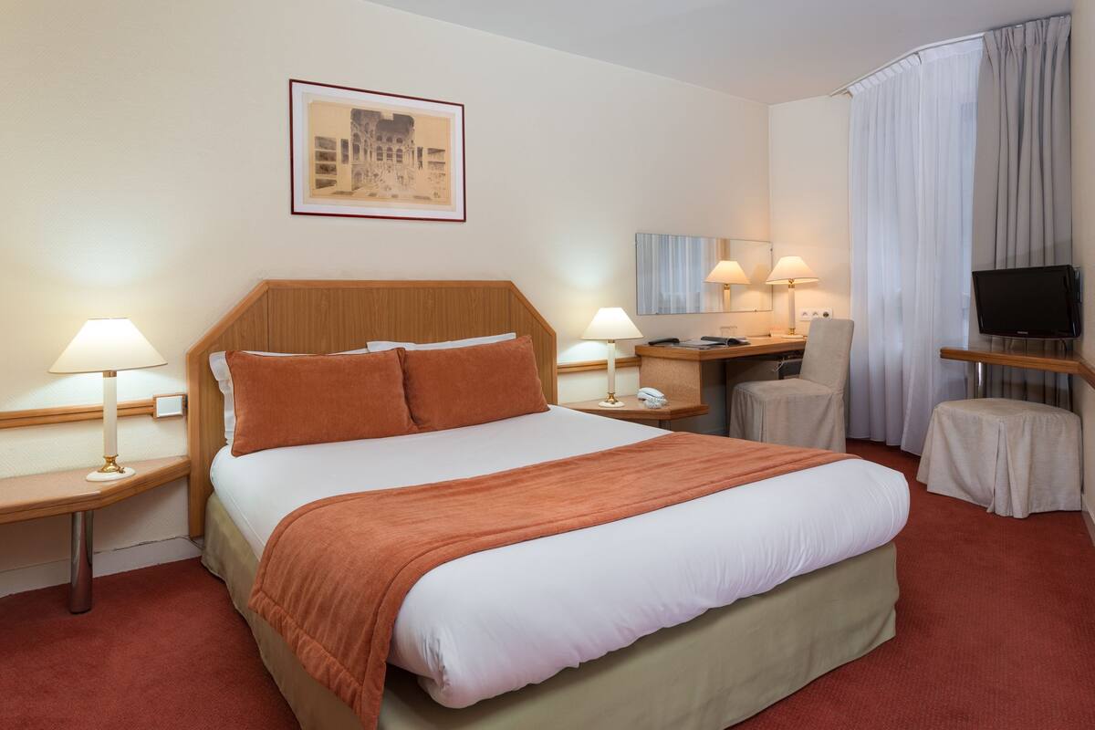 A hotel room with a large bed, two bedside tables with lamps, a desk with a chair, a television, and a framed picture on the wall.