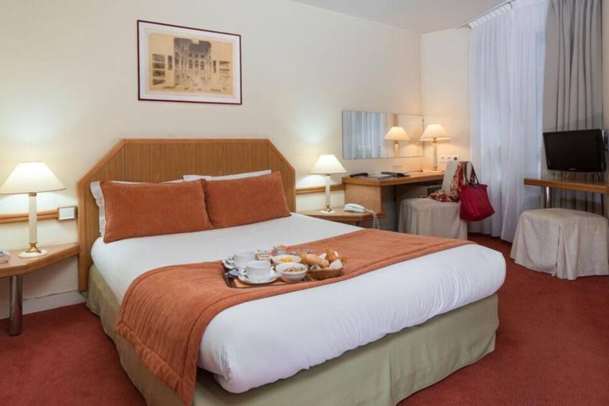 A hotel room with a bed, two bedside tables with lamps, a television, a small table with a red bag, and a framed picture on the wall.
