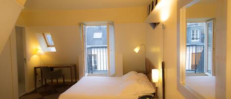 A hotel room with a bed, a desk, a chair, a lamp, and a window.