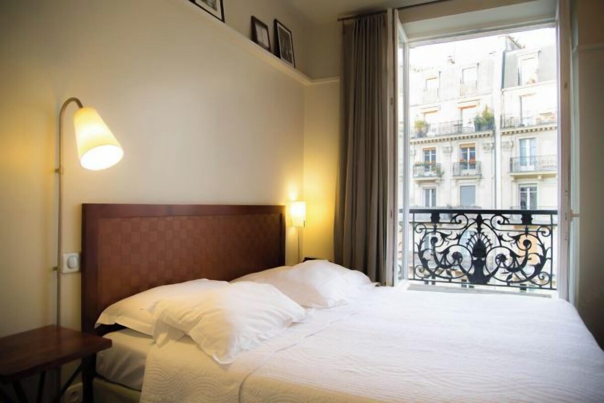 Une chambre d’hôtel avec un lit, deux lampes de chevet, une table de chevet et une vue sur les bâtiments, visible par une fenêtre donnant sur un balcon avec une rambarde.