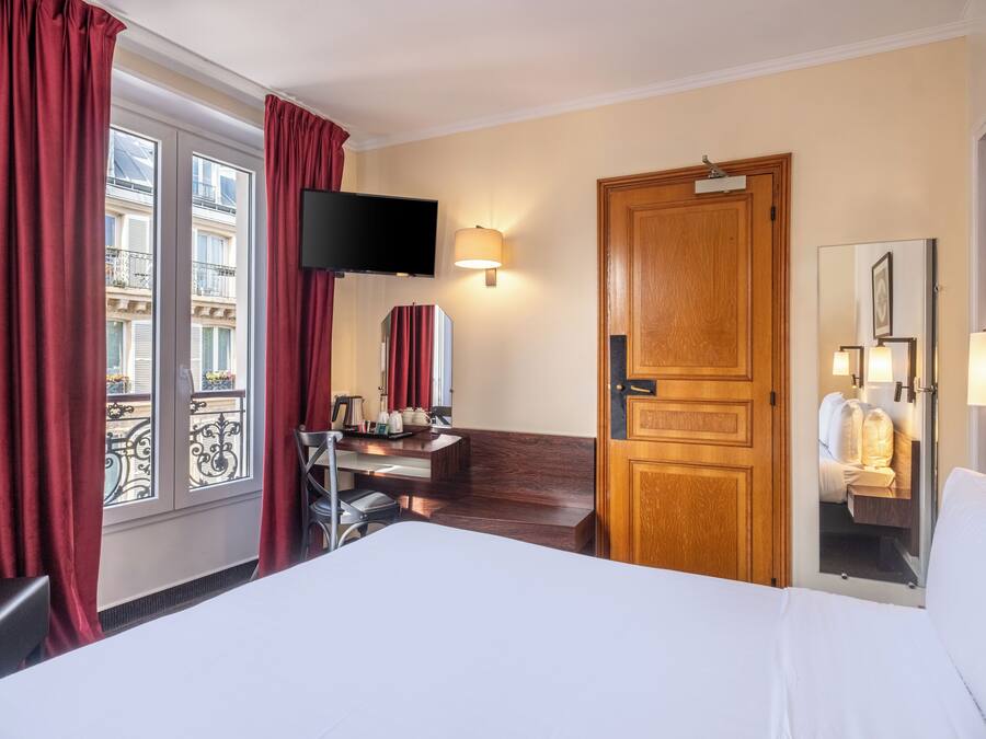 Una habitació d'hotel amb un llit, una taula de treball amb una cadira, un televisor fixat a la paret, una porta de fusta i una finestra amb cortines.
