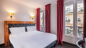 A hotel room with a large bed, two wall-mounted lamps, and a view of the city through large windows with red curtains.