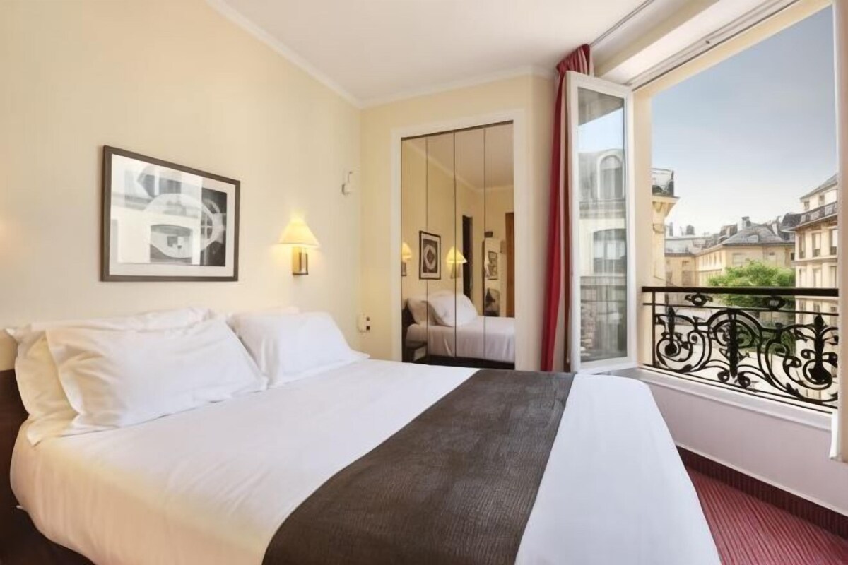 A hotel room with a bed, a window with a view of buildings, a door, and a framed picture on the wall.