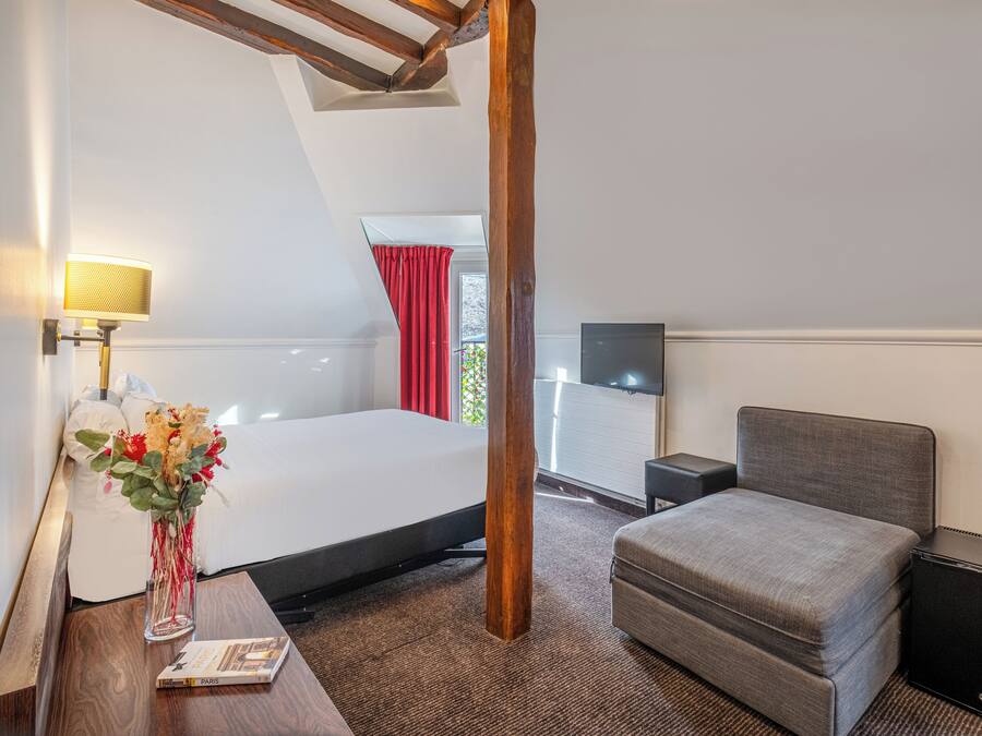 A hotel room with a bed, a desk with a lamp, a small table with a vase of flowers, a grey sofa, and a TV on a stand.