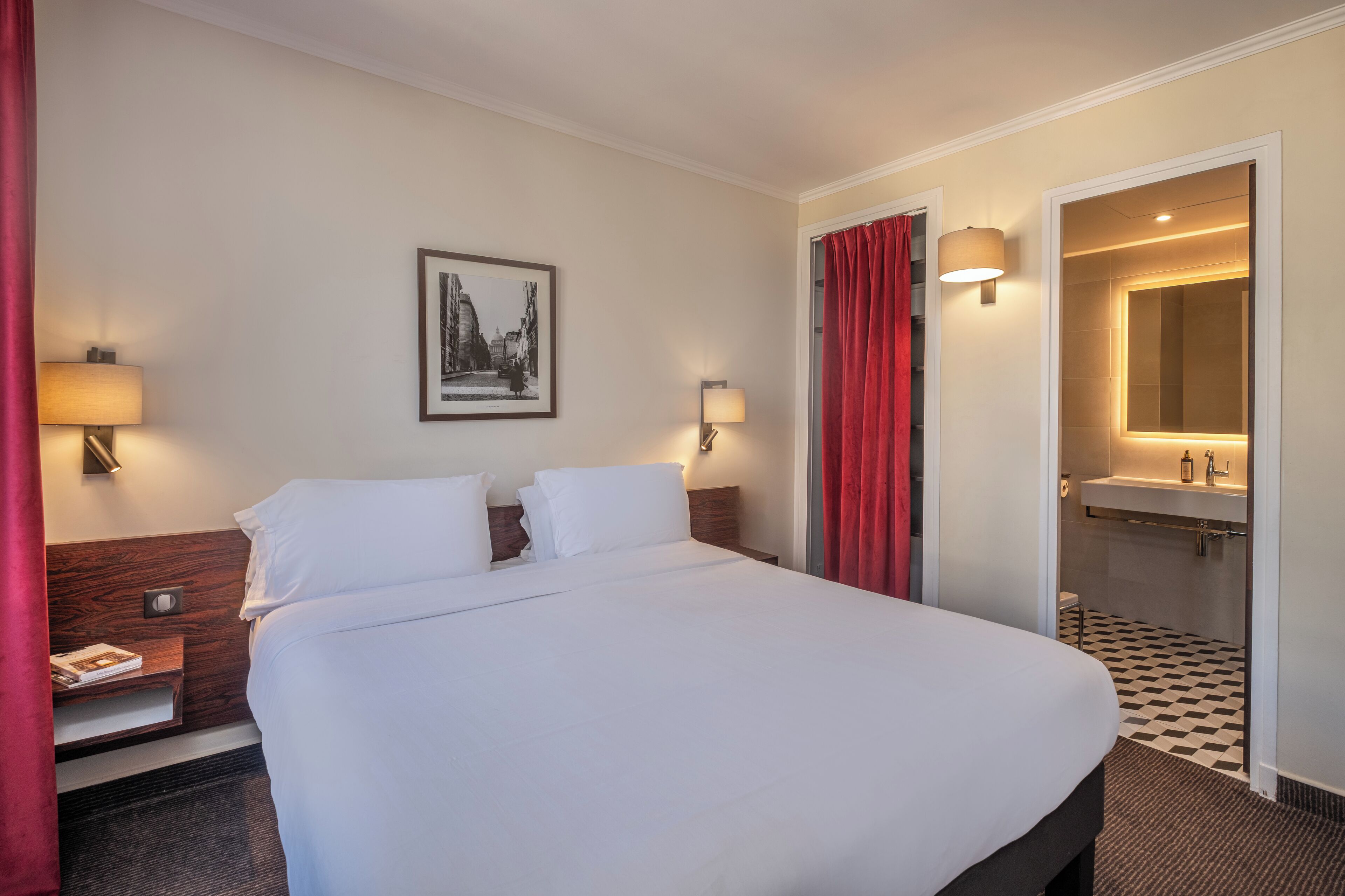 Una habitación de hotel con una cama grande, dos mesitas de noche con lámparas, un cuadro en la pared, una cortina roja y un baño visible a través de una puerta abierta.