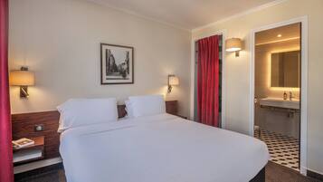 Una habitación de hotel con una cama grande, dos mesitas de noche con lámparas, un cuadro en la pared, una cortina roja y un baño visible a través de una puerta abierta.