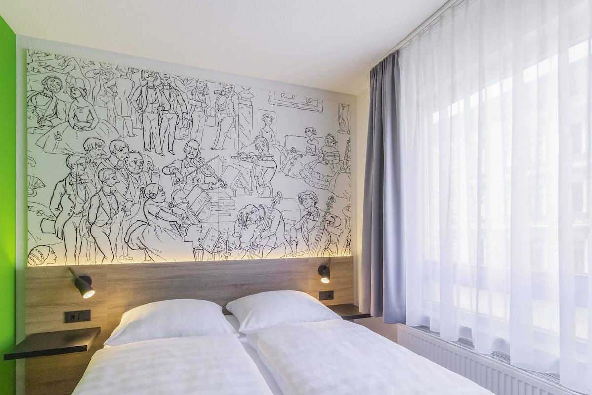 A hotel room with a large wall mural, a bed with white linens, a nightstand, and a window with sheer curtains.