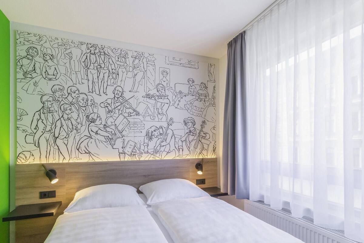 A hotel room with a large wall mural, a bed with white linens, a nightstand, and a window with sheer curtains.