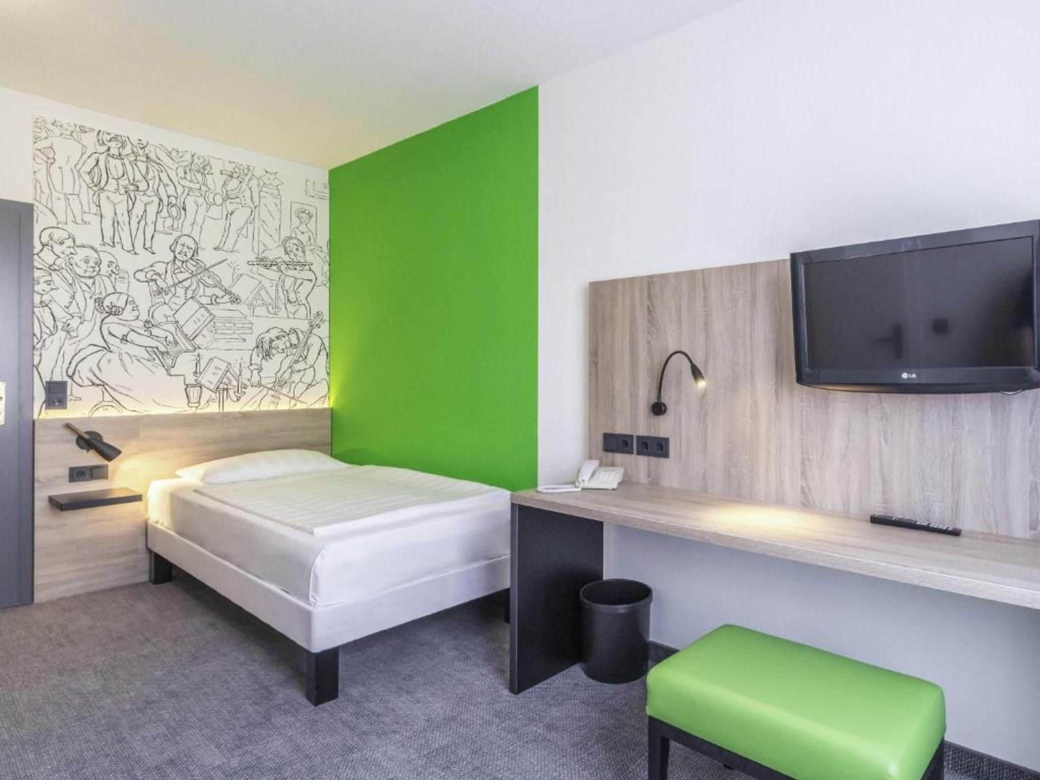 Habitación de hotel con cama, escritorio con silla, televisión y una pared verde decorativa.