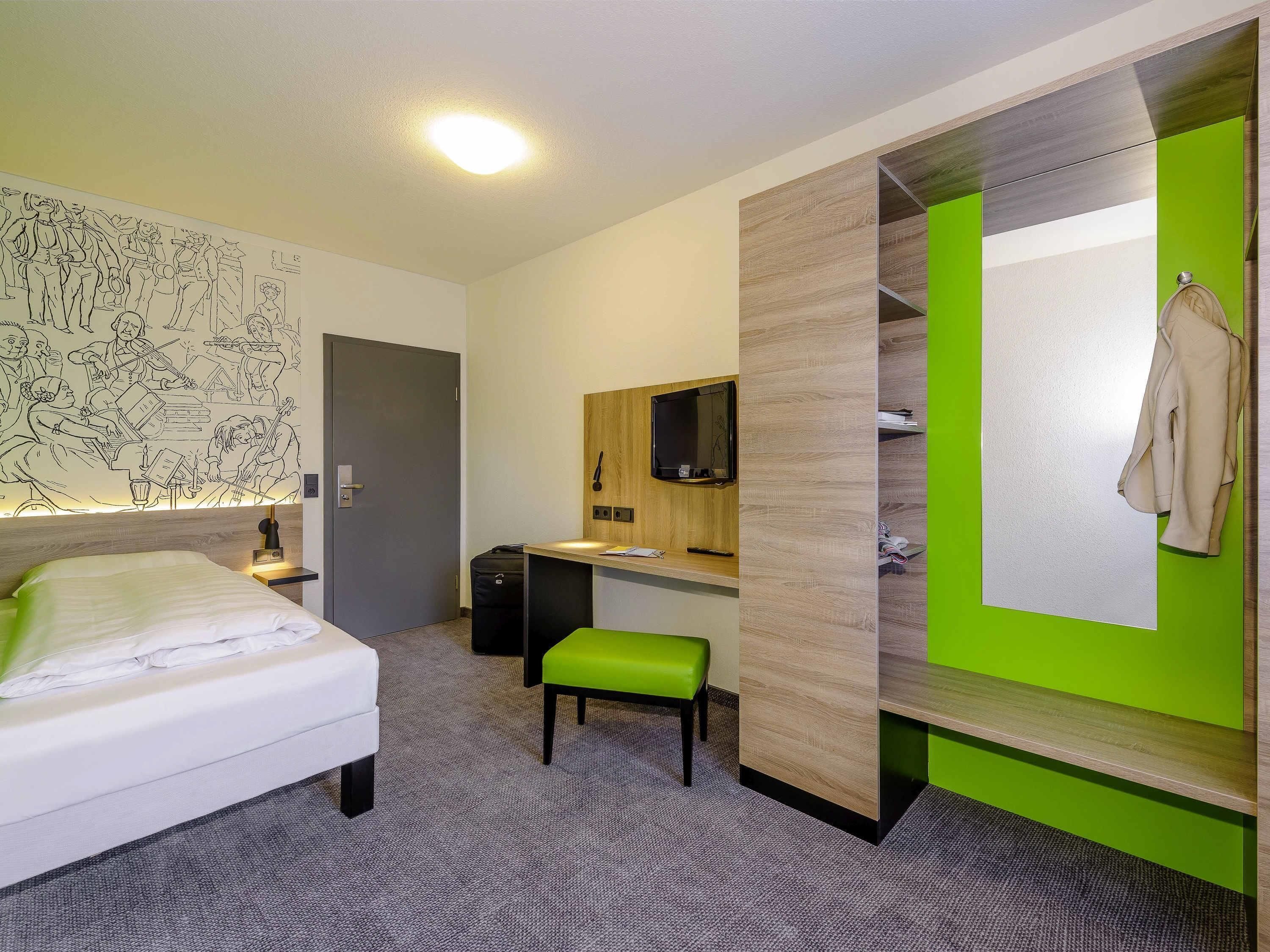 A hotel room with a bed, a desk, a green chair, a TV, and a wall with a mural.