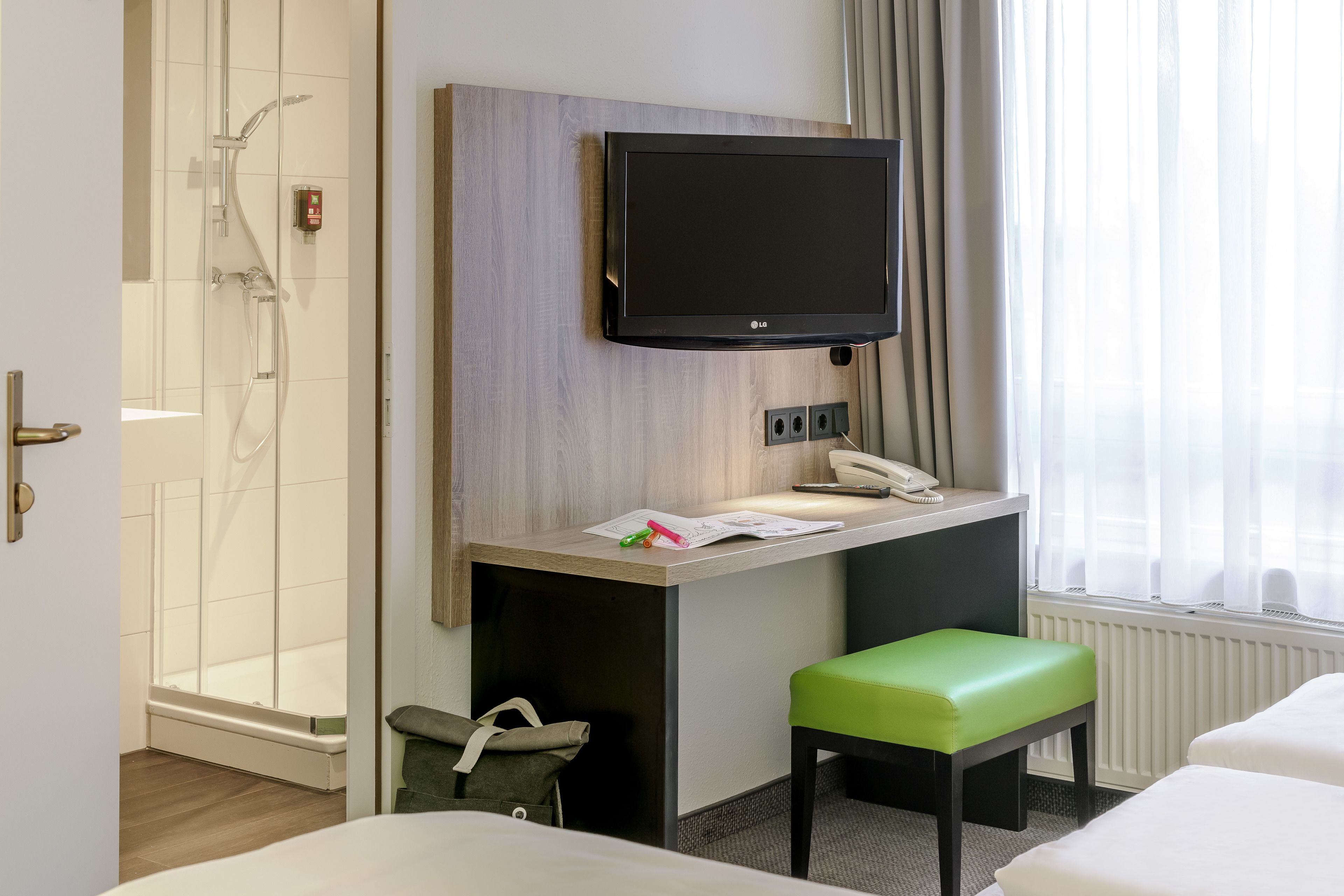 A hotel room with a shower, a desk with a flat-screen TV, a telephone, and a green bench.