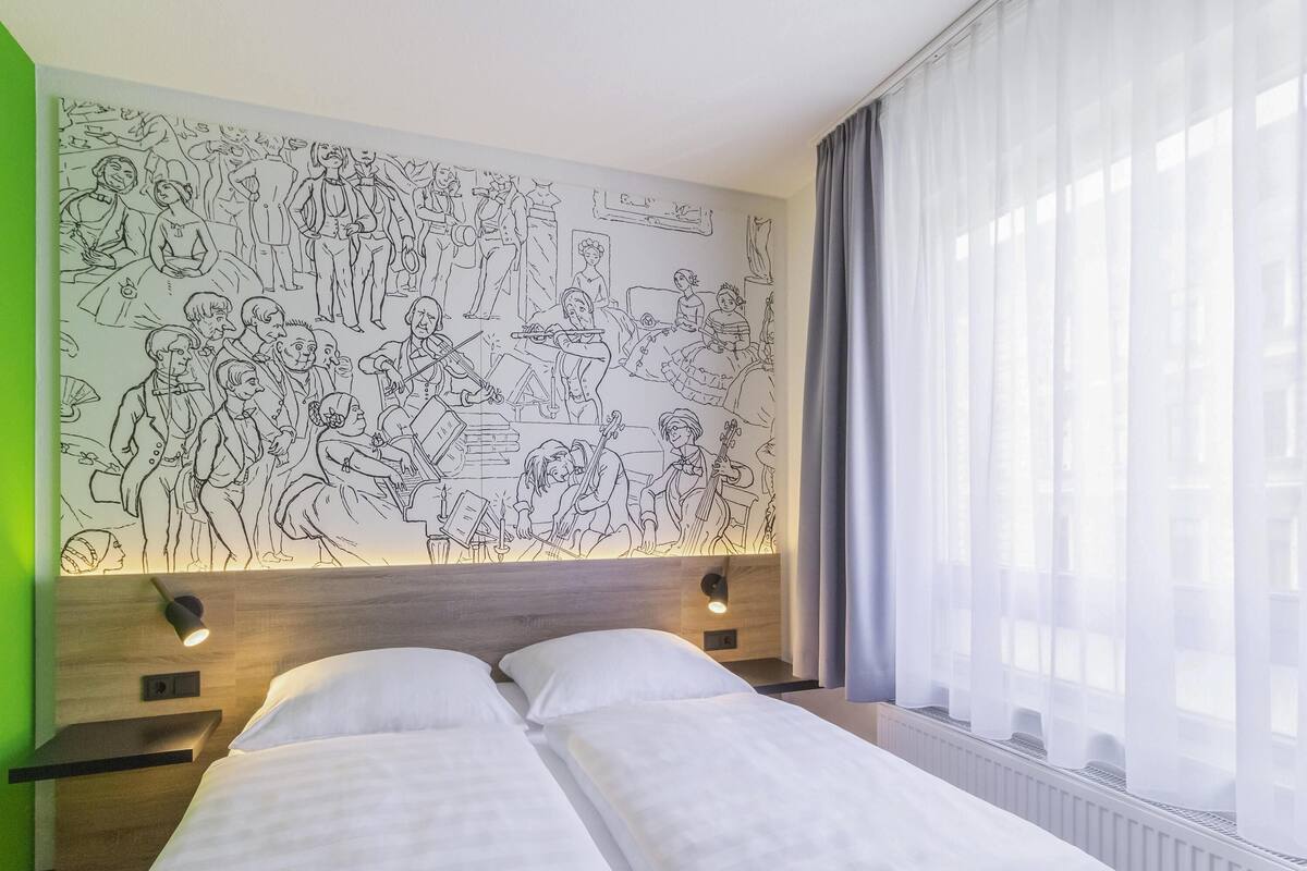 A hotel room with a large wall mural, a bed with white linens, a nightstand, and a window with sheer curtains.