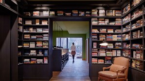 Eine Bibliothek mit Bücherregalen an beiden Seiten eines zentralen Ganges, einem Ledersessel und einer Person, die auf ein Fenster zugeht.