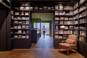 Una biblioteca con estanterías a ambos lados de un pasillo central, un sillón de cuero y una persona caminando hacia una ventana.