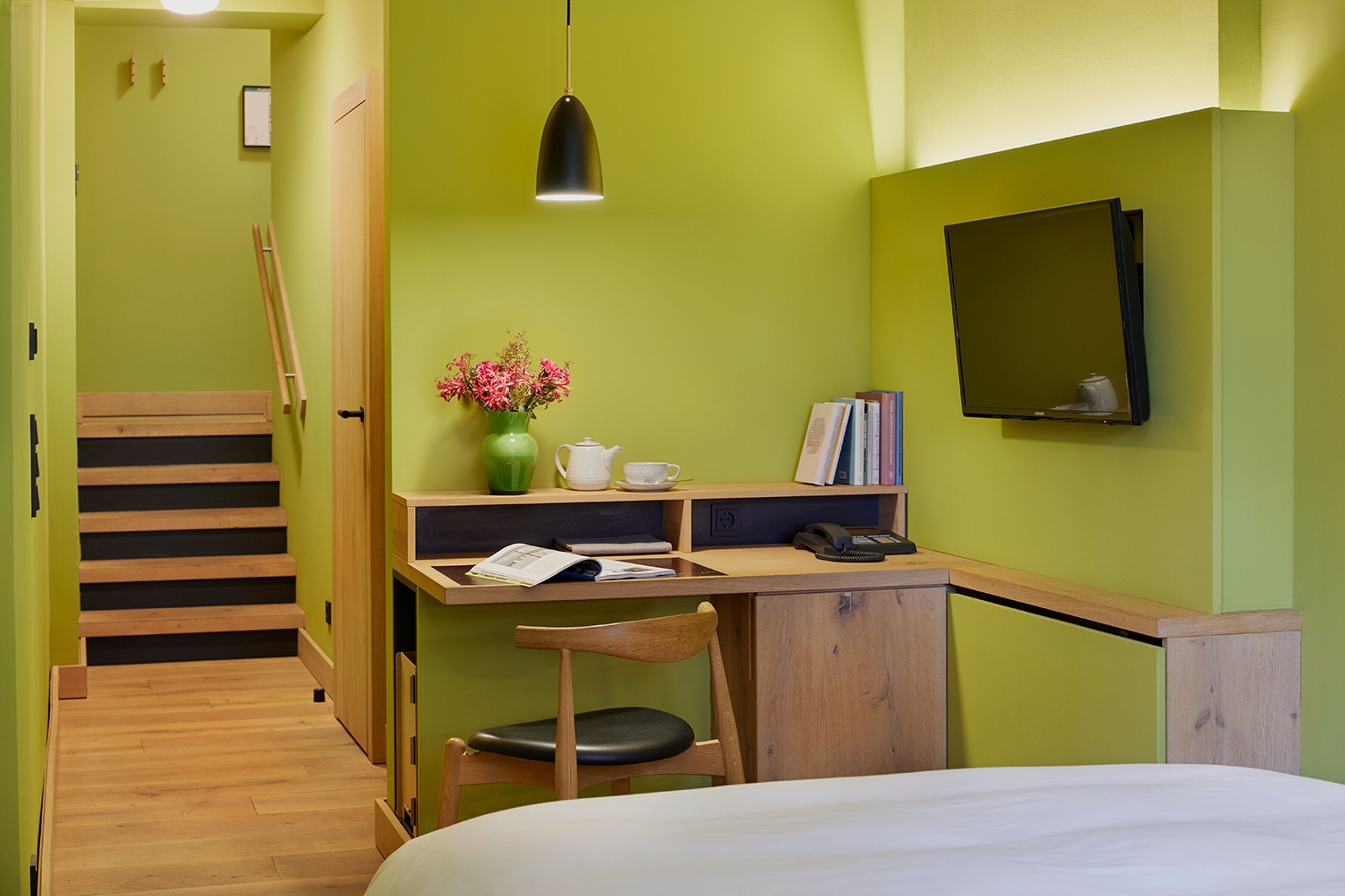 A compact room with a bed, a desk with a chair, a small table with a vase of flowers, a bookshelf, and a mirror on the wall.