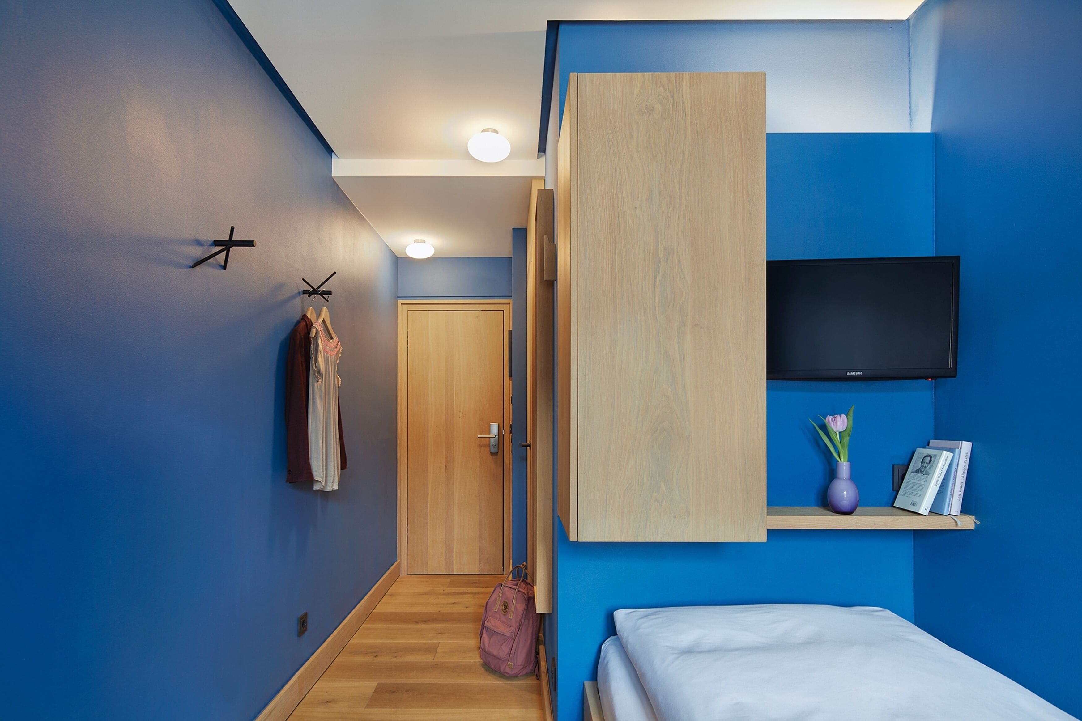 Un dormitorio moderno con una cama, un televisor montado en la pared, un armario de madera y un pasillo que conduce a una puerta.