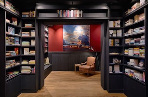 Una biblioteca con estanterías a ambos lados, suelo de madera, pared roja con un cuadro y una silla en el centro.