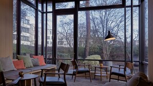 Ein gemütlicher Sitzbereich mit einer Couch, Sesseln und Tischen, große Fenster mit Blick auf Bäume und Gebäude.