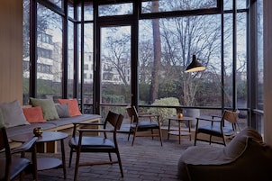 Cómoda zona de estar con sofá, sillas y mesas, ventanas amplias con vistas a árboles y edificios.