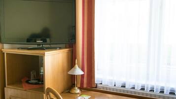 A hotel room with a television on a wooden cabinet, a table with a lamp, and a window with sheer curtains.