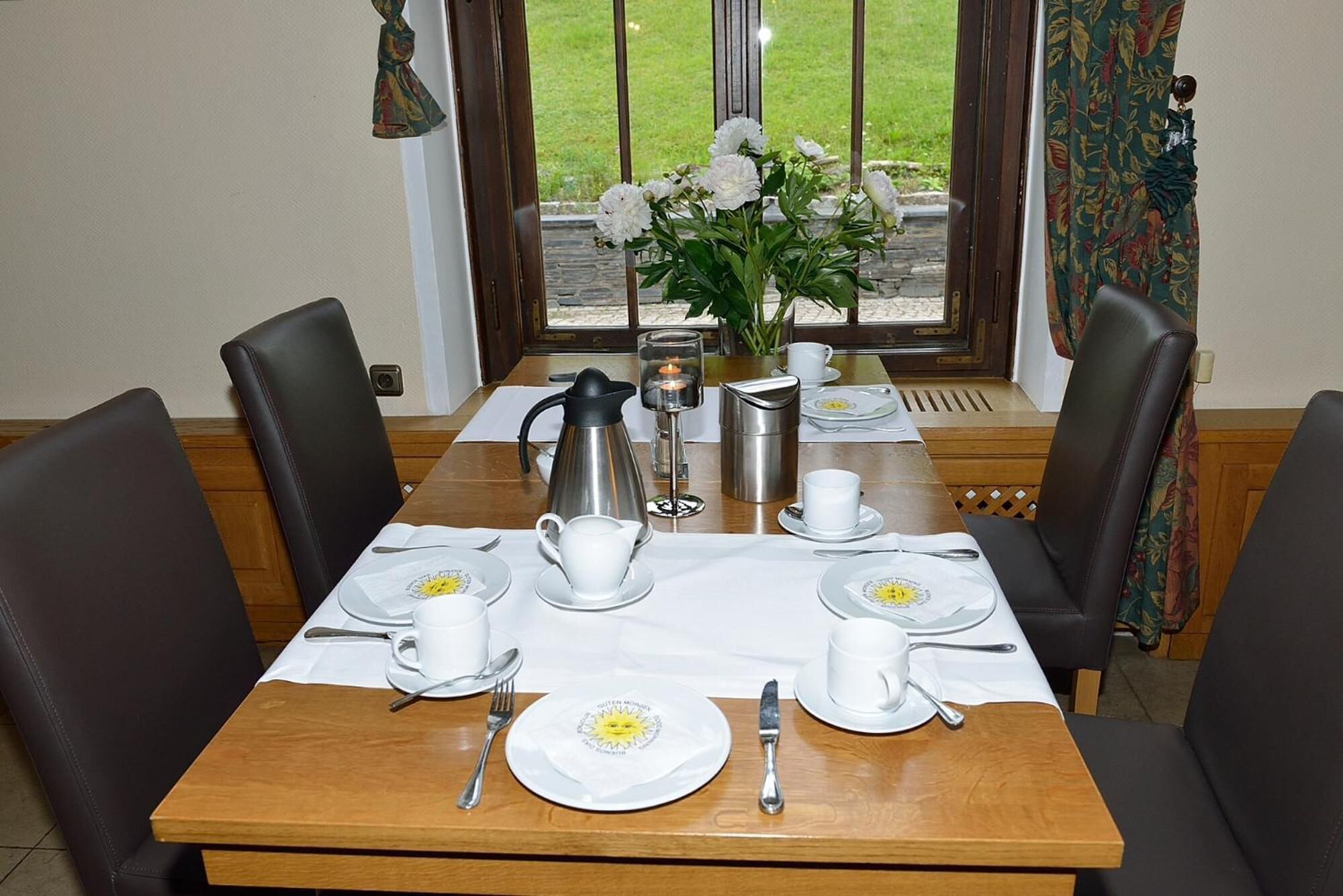 A dining table set for four with white plates, cups, and cutlery, a coffee pot, and a candle.