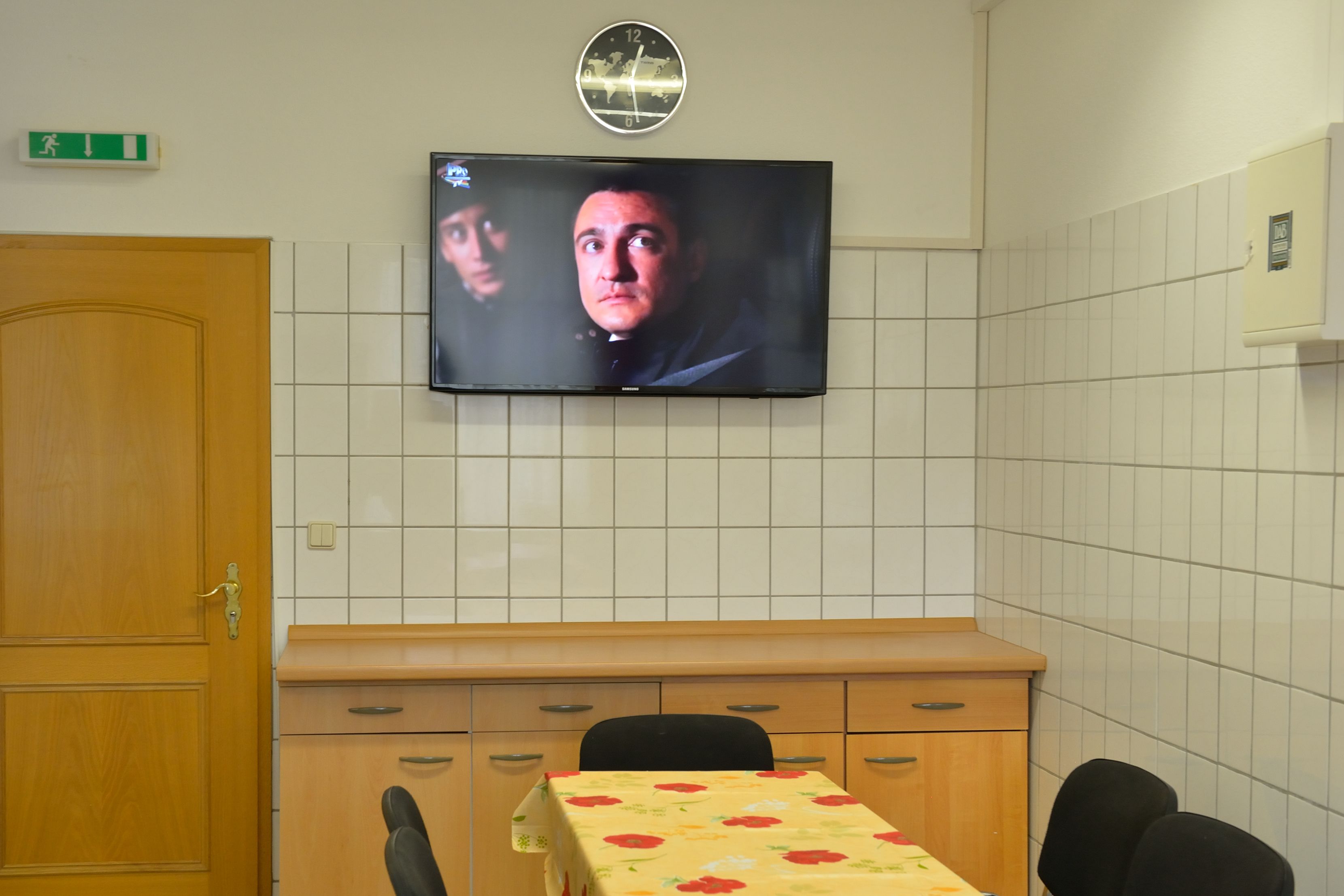 Una habitació amb televisió penjada a la paret, una porta de fusta, un mur amb rajoles, una taula amb mantell de flors i una cadira.
