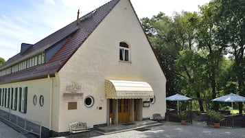 Un edificio tradicional con techo inclinado, entrada principal y un letrero que dice “BELAIR RESTAURANT”. Hay áreas de estar al aire libre con sombrillas y plantas en macetas.