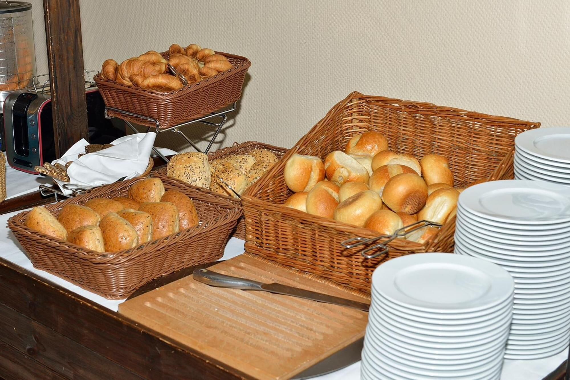 A buffet table with various types of bread and rolls in wicker baskets, accompanied by stacks of white plates.