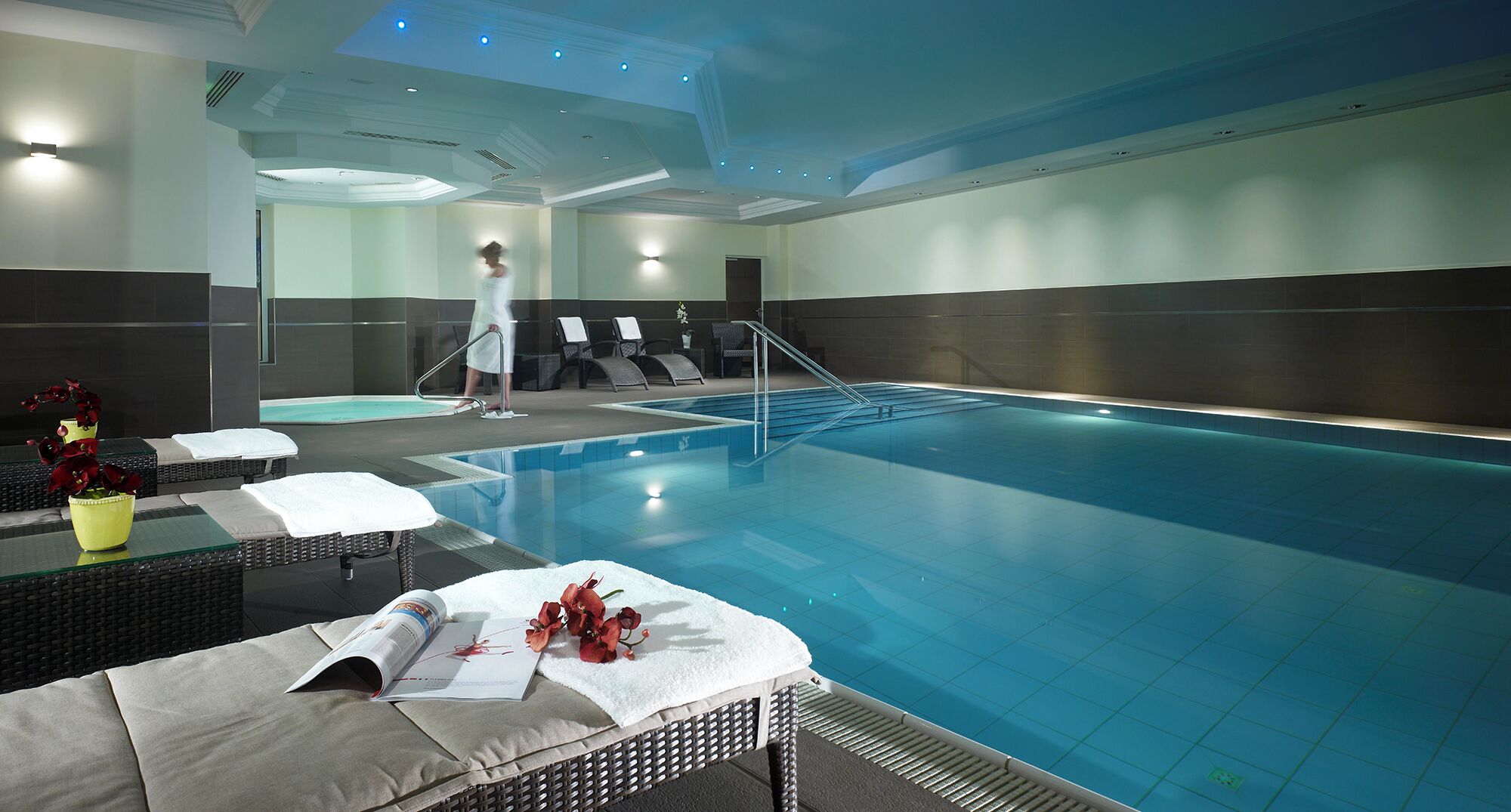 A swimming pool with lounge chairs and towels, a person walking by, and a ceiling with recessed lighting.