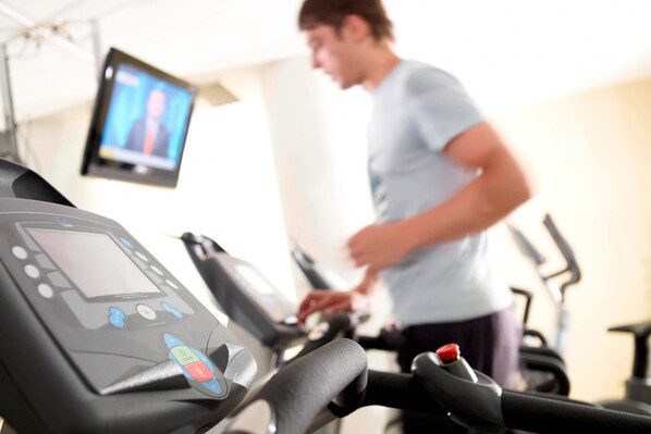 A treadmill with a digital display and control panel, a television mounted on the wall, and a person using the treadmill.