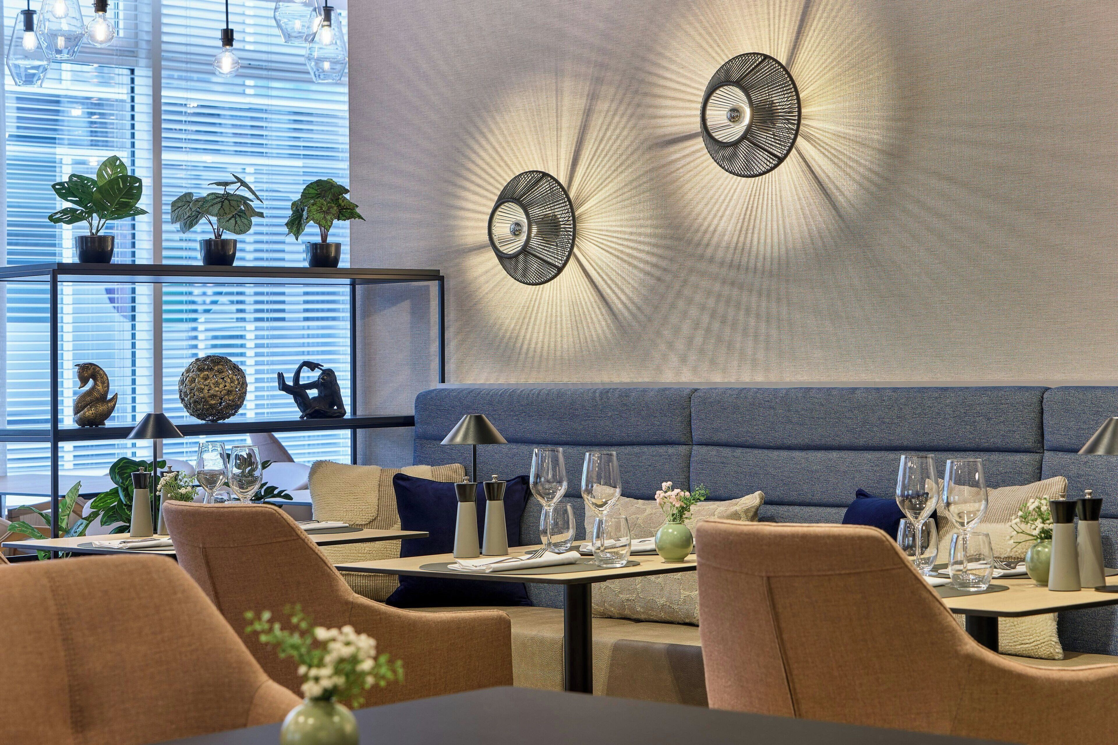 Un comedor moderno con un banco tapizado azul, varias mesas con sillas y apliques decorativos en la pared.