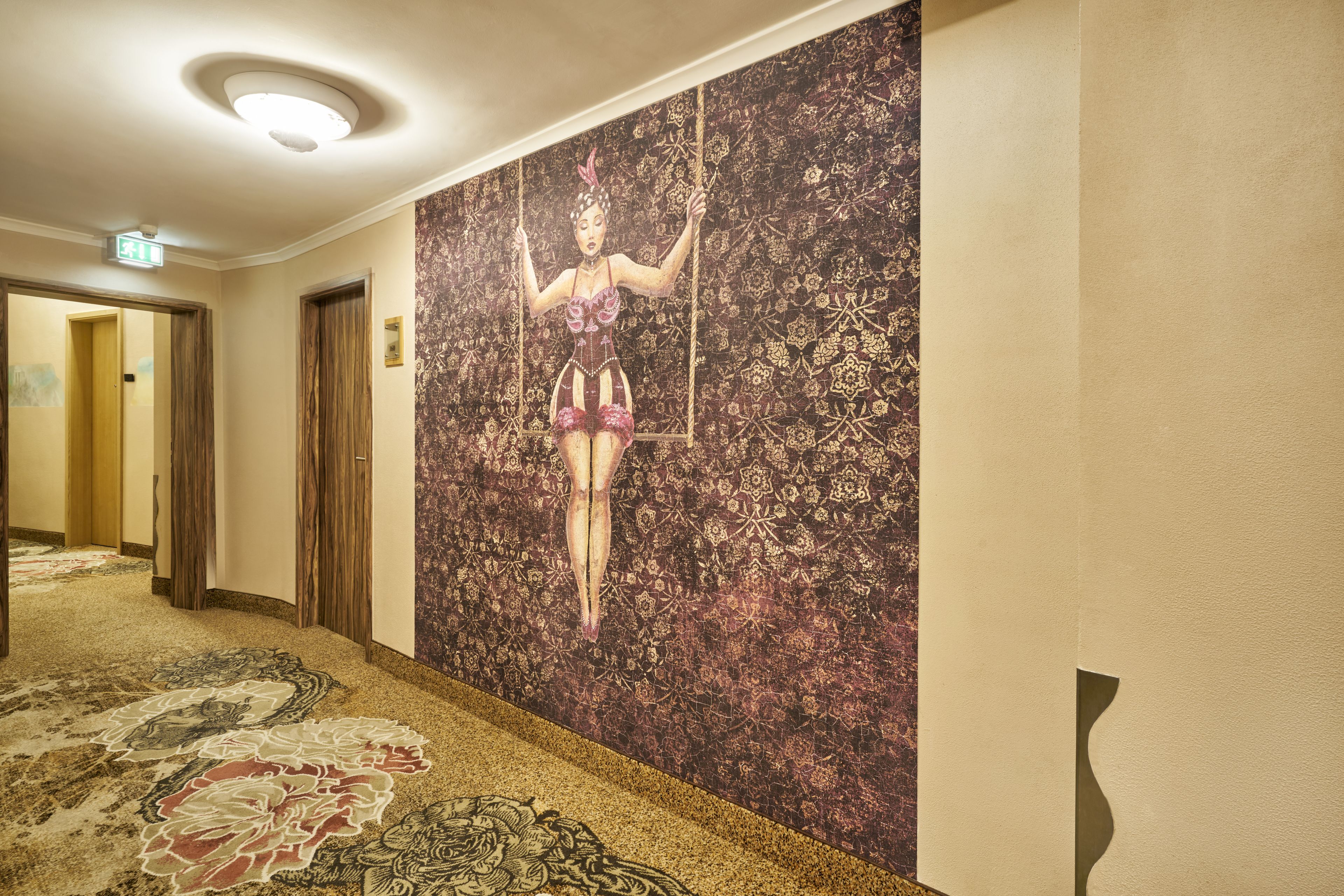 Un couloir orné d’une grande fresque représentant une femme sur une balançoire, doté de revêtements de sol en moquette et de portes donnant accès à d’autres pièces.