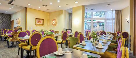 A hotel dining area with multiple tables and chairs, a patterned wall, and a large window.