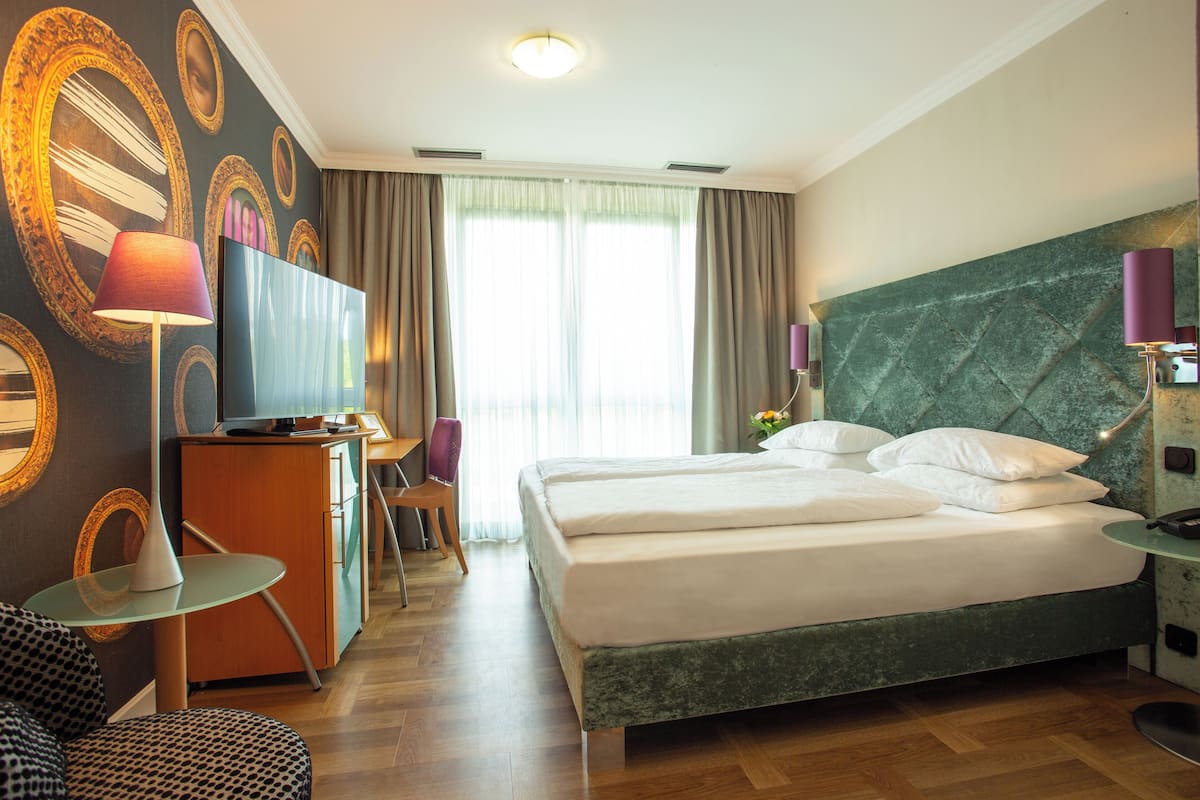 A hotel room with a large bed, a desk with a TV, a chair, and a small table.