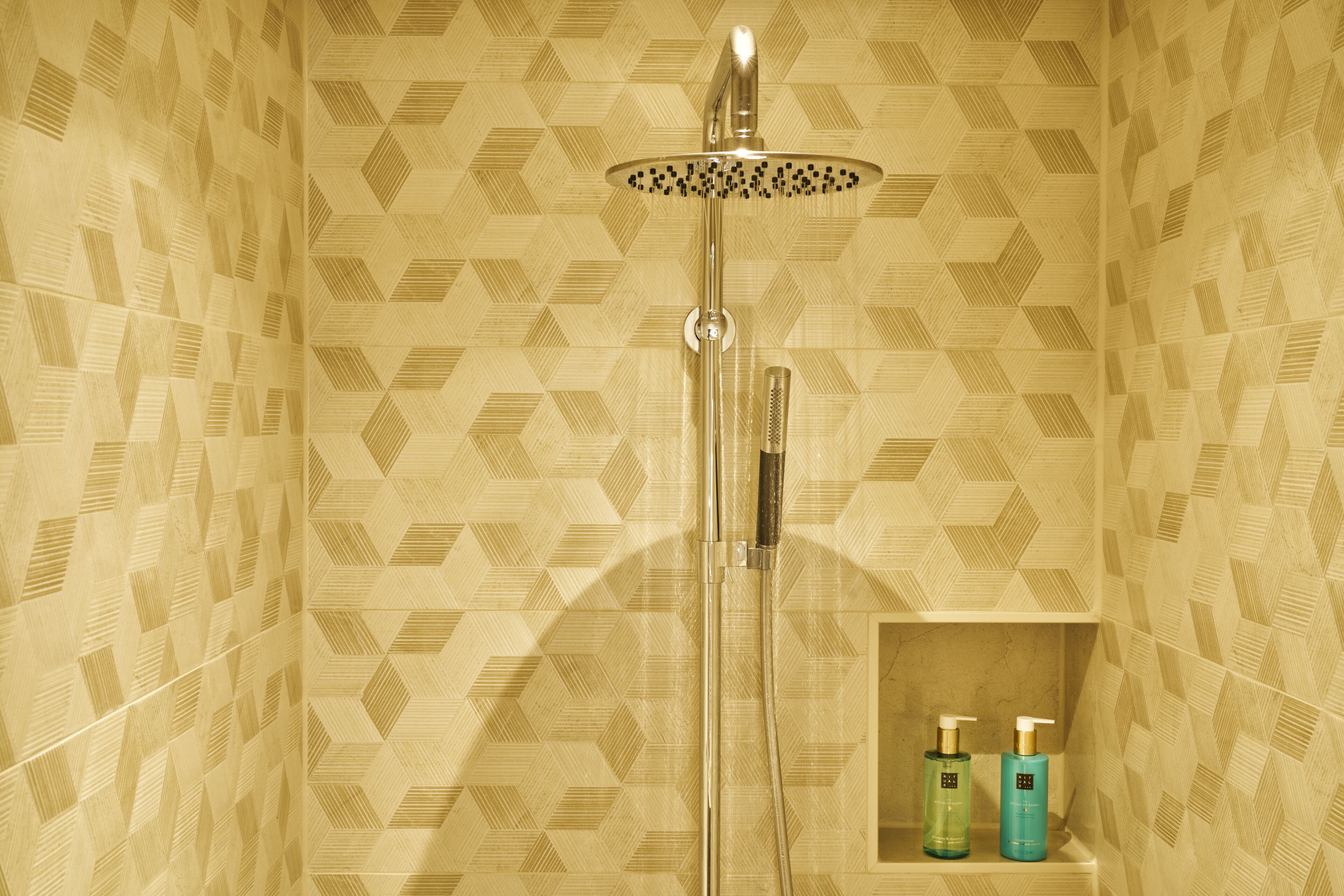 A shower with a geometric patterned wall, a handheld showerhead, and two bottles of shampoo on a shelf.