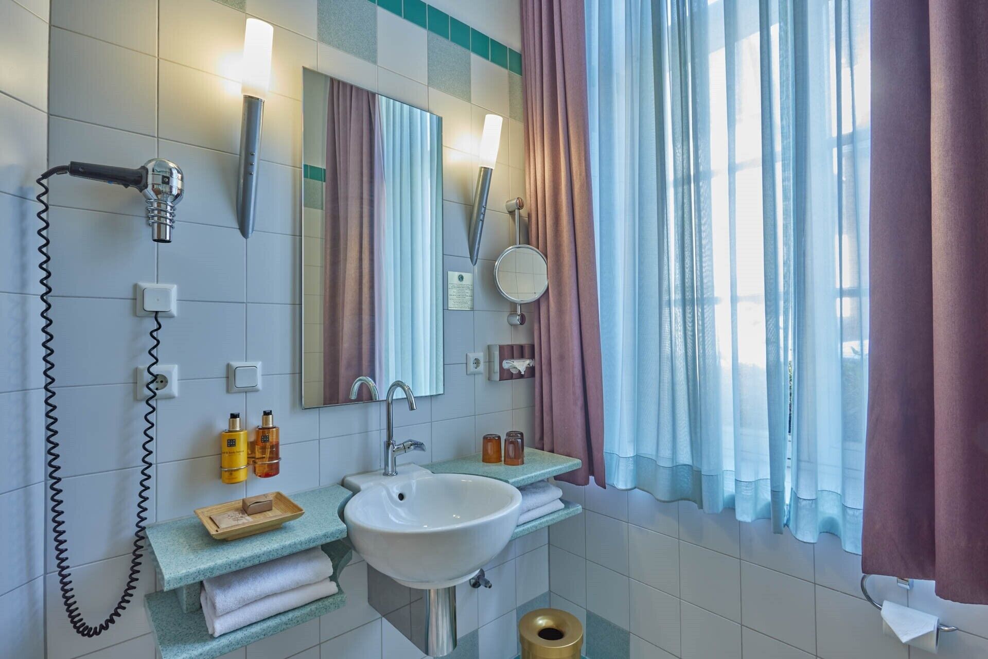 Ein Badezimmer mit Waschbecken, Spiegel und Handtuchhalter. Ein Föhn ist an der Wand angebracht und ein Fenster mit Vorhängen vorhanden.
