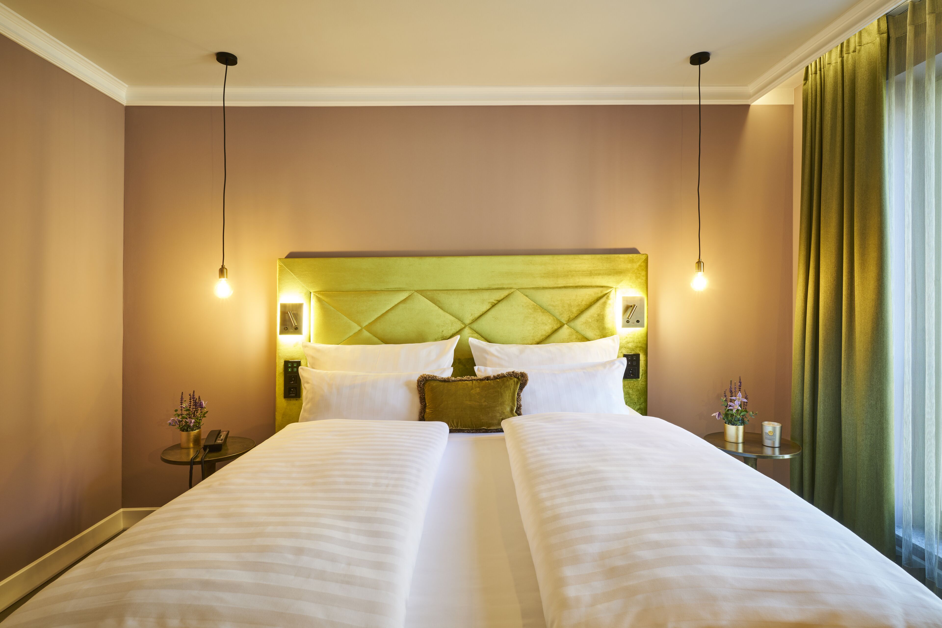 A hotel room with a large bed, two bedside tables, a green headboard, and a view of the outdoors through a window with curtains.
