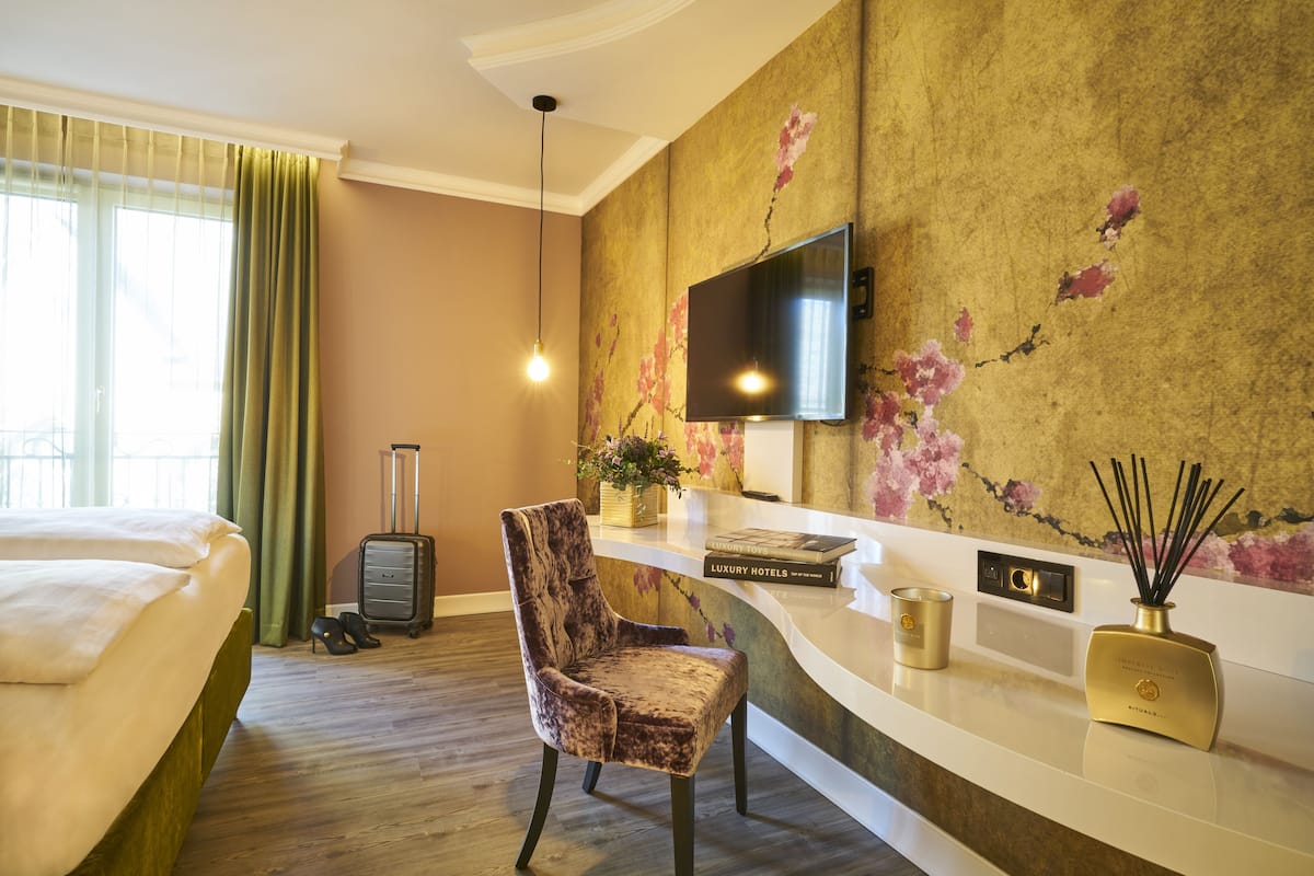 A hotel room with a bed, a chair, a desk, a television, and a wall with floral wallpaper.