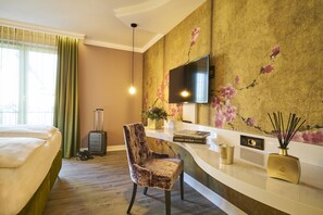 Quarto de hotel com cama, poltrona, escrivaninha, televisão e parede com papel de parede floral.