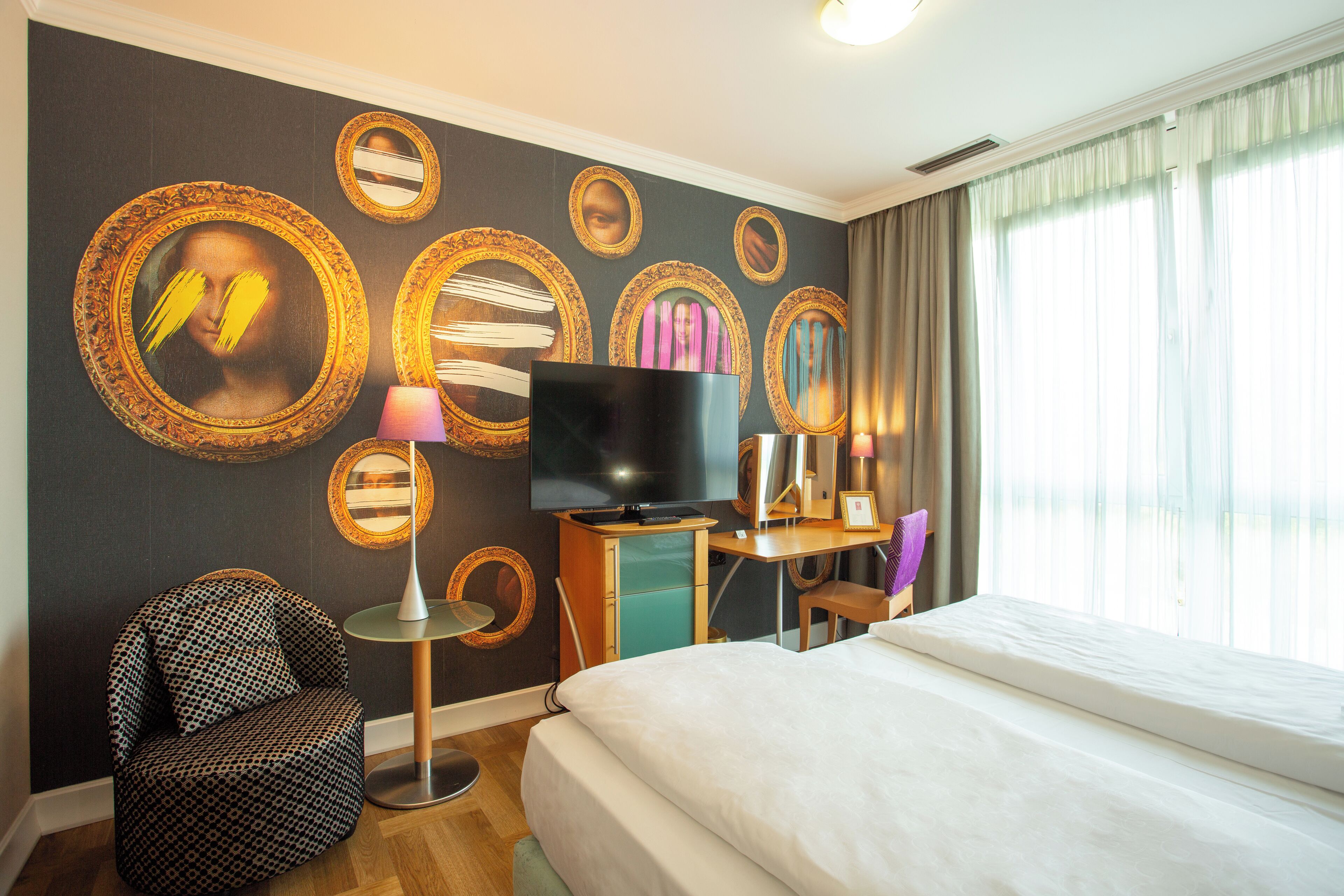 Habitación de hotel con cama, televisor, escritorio, silla y varios espejos enmarcados en la pared.